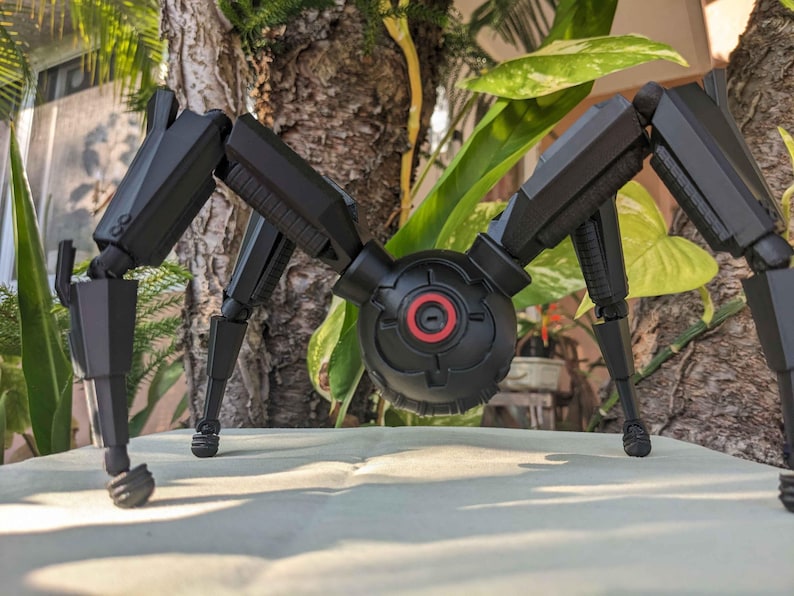 Pode incluir: Um modelo preto, mec&acirc;nico, semelhante a uma aranha, com pernas articuladas e um corpo central. O corpo apresenta um design circular com um detalhe vermelho. O modelo est&aacute; sobre um fundo natural com folhagem.