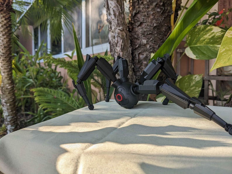 Pode incluir: Um brinquedo de aranha mec&acirc;nica preta com pernas articuladas e um corpo redondo. O brinquedo tem um detalhe circular vermelho no corpo. A aranha est&aacute; posicionada em uma superf&iacute;cie de cor clara, com vegeta&ccedil;&atilde;o ao fundo.
