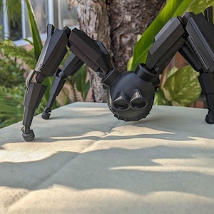 Pode incluir: Um brinquedo de rob&ocirc; aranha preto articulado com v&aacute;rias pernas e um corpo redondo. O brinquedo &eacute; feito de pl&aacute;stico e tem um acabamento fosco. A aranha est&aacute; pousada em uma superf&iacute;cie de cor clara, com vegeta&ccedil;&atilde;o ao fundo.
