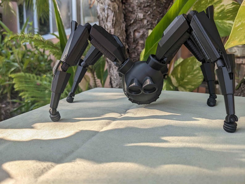 Pode incluir: Um brinquedo de rob&ocirc; aranha preto articulado com v&aacute;rias pernas e um corpo redondo. O brinquedo &eacute; feito de pl&aacute;stico e tem um acabamento fosco. A aranha est&aacute; pousada em uma superf&iacute;cie de cor clara, com vegeta&ccedil;&atilde;o ao fundo.