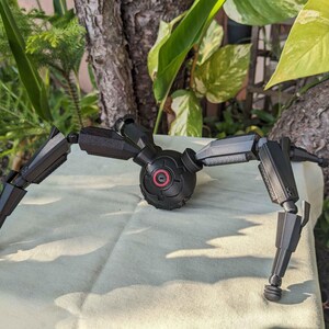 Pode incluir: Um modelo mec&acirc;nico preto, semelhante a uma aranha, com pernas articuladas e um corpo central. O modelo tem um detalhe circular vermelho no corpo. As pernas s&atilde;o segmentadas e terminam em p&eacute;s arredondados.