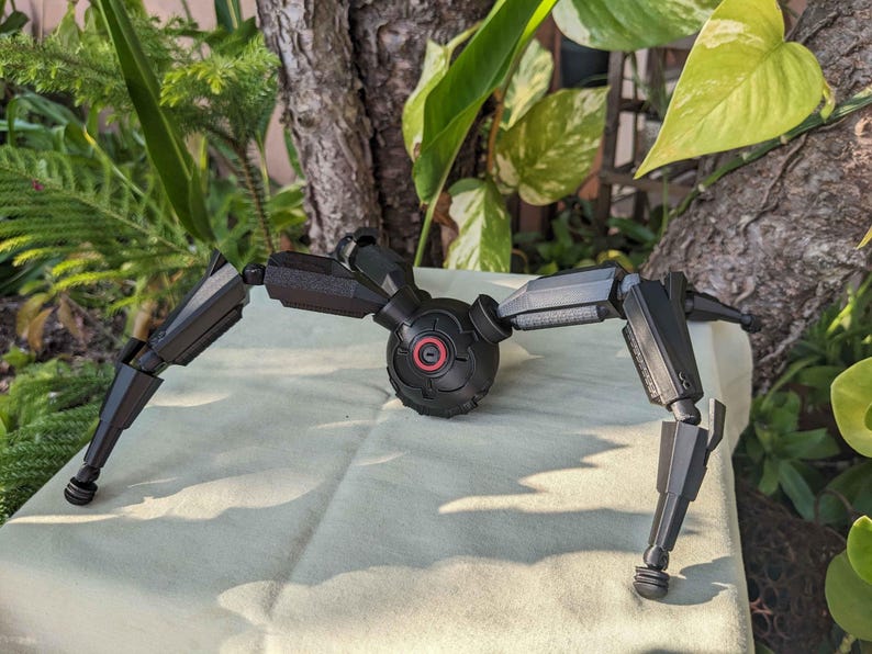 Pode incluir: Um modelo mec&acirc;nico preto, semelhante a uma aranha, com pernas articuladas e um corpo central. O modelo tem um detalhe circular vermelho no corpo. As pernas s&atilde;o segmentadas e terminam em p&eacute;s arredondados.