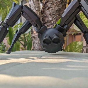 Pode incluir: Um brinquedo de aranha preto articulado, com v&aacute;rias pernas articuladas e um corpo arredondado. O brinquedo tem um design detalhado, com componentes mec&acirc;nicos vis&iacute;veis e uma superf&iacute;cie texturizada. A aranha est&aacute; pousada numa superf&iacute;cie de cor clara, com um fundo desfocado de vegeta&ccedil;&atilde;o.