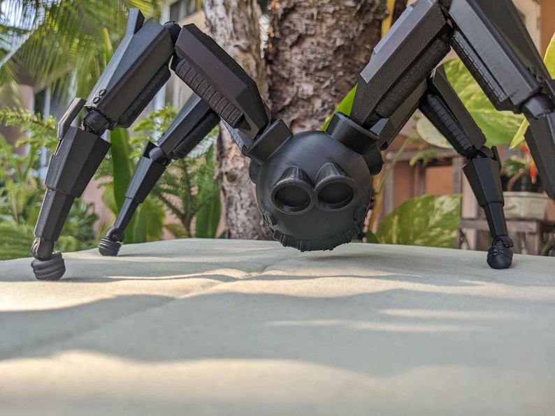 Pode incluir: Um brinquedo de aranha preto articulado, com v&aacute;rias pernas articuladas e um corpo arredondado. O brinquedo tem um design detalhado, com componentes mec&acirc;nicos vis&iacute;veis e uma superf&iacute;cie texturizada. A aranha est&aacute; pousada numa superf&iacute;cie de cor clara, com um fundo desfocado de vegeta&ccedil;&atilde;o.