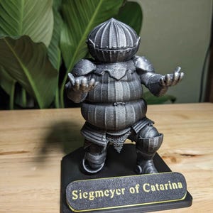 May include: A metallic gray figurine of a knight in full armor, posed with open hands. The figurine is mounted on a black base with the text "Siegmeyer of Catarina" in gold lettering. The knight's helmet and armor are detailed.