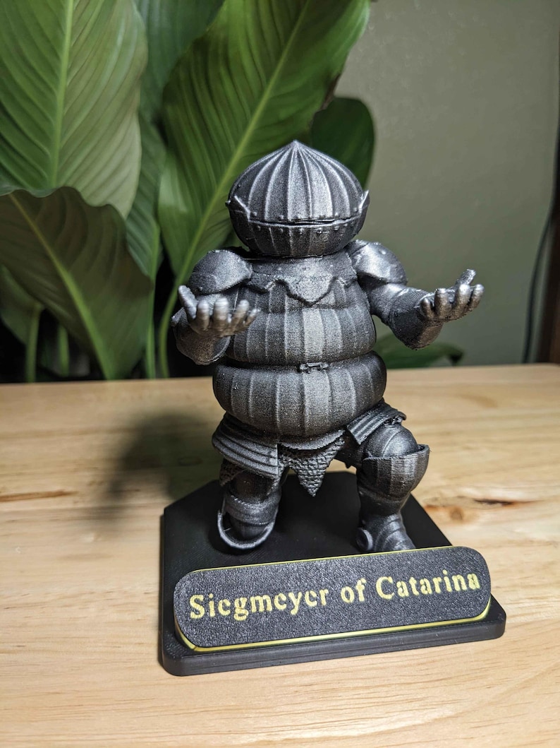 May include: A metallic gray figurine of a knight in full armor, posed with open hands. The figurine is mounted on a black base with the text "Siegmeyer of Catarina" in gold lettering. The knight's helmet and armor are detailed.