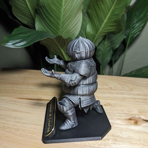 May include: A silver-colored figurine of a kneeling knight with outstretched arms. The knight is wearing armor and a helmet. The figurine is on a black base with gold lettering that reads "Player of Catarina".