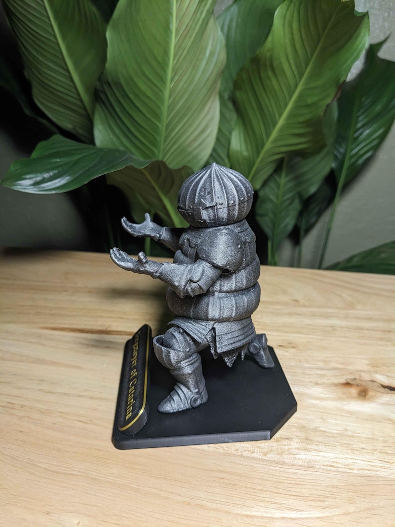 May include: A silver-colored figurine of a kneeling knight with outstretched arms. The knight is wearing armor and a helmet. The figurine is on a black base with gold lettering that reads "Player of Catarina".