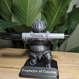 Puede incluir: Una figura gris metálica de un caballero sosteniendo un marcador Sharpie. La figura está sobre una base negra con el texto "Siegmeyer of Catarina" en letras doradas. El Sharpie es blanco y negro.