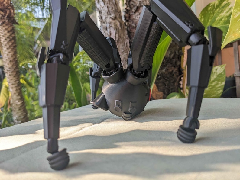 Pode incluir: Um brinquedo de aranha rob&oacute;tica preta articulada com v&aacute;rias pernas e uma cabe&ccedil;a com dois detalhes circulares semelhantes a olhos. O brinquedo est&aacute; posicionado em uma superf&iacute;cie de cor clara, com um fundo verde desfocado de folhagem.