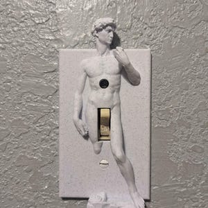 May include: A light gray light switch cover featuring a sculpted relief of Michelangelo's David. The switch is in the center, with the word "ON" visible. The cover is mounted on a textured gray wall.