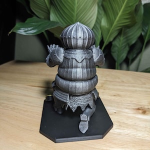 May include: A gray 3D-printed figurine of a knight in armor, kneeling on a black hexagonal base. The knight's helmet and armor have detailed textures. The background features large green leaves.