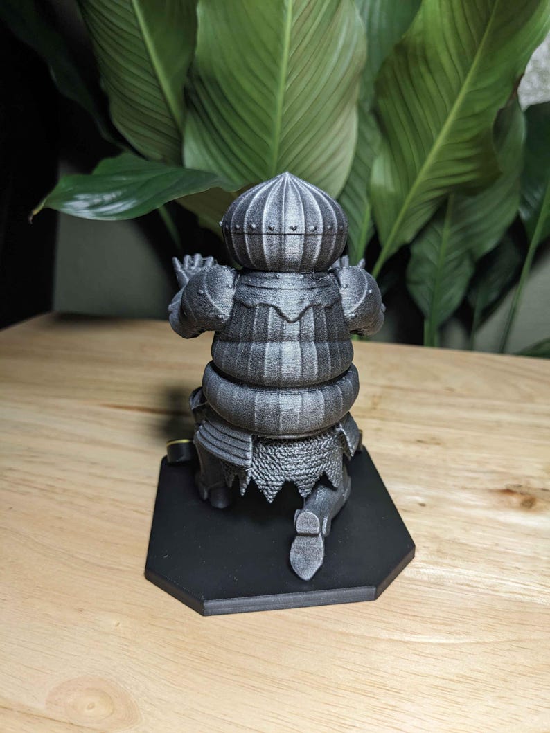 May include: A gray 3D-printed figurine of a knight in armor, kneeling on a black hexagonal base. The knight's helmet and armor have detailed textures. The background features large green leaves.