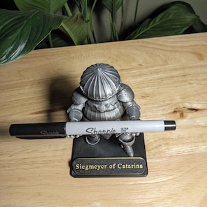 May include: A silver-colored figurine of a knight, holding a black and silver Sharpie marker. The figurine is on a black base with the text "Siegmeyer of Catarina". The background includes green leaves and a wooden surface.