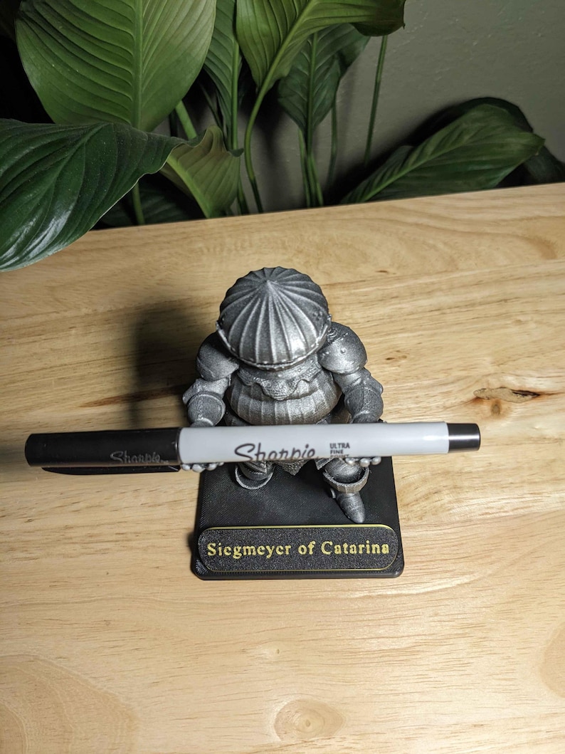 May include: A silver-colored figurine of a knight, holding a black and silver Sharpie marker. The figurine is on a black base with the text "Siegmeyer of Catarina". The background includes green leaves and a wooden surface.