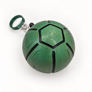 Può includere: Un portachiavi sferico verde con linee nere incassate che formano un motivo geometrico. L'oggetto presenta un esagono centrale ed è fissato a un anello circolare verde tramite una piccola clip metallica e una singola perlina nera.