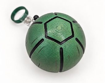 May include: A spherical green keychain with black recessed lines forming a geometric pattern. The object features a central hexagon and is attached to a green circular ring by a small metal clip and a single black bead, creating a compact accessory.