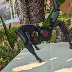Pode incluir: Um modelo preto e mec&acirc;nico semelhante a uma aranha, com pernas articuladas e um corpo central. O modelo tem um componente circular com um centro vermelho. O fundo inclui vegeta&ccedil;&atilde;o e um tronco de &aacute;rvore.
