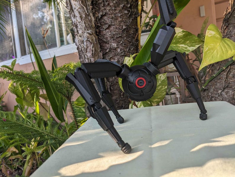 Pode incluir: Um modelo preto e mec&acirc;nico semelhante a uma aranha, com pernas articuladas e um corpo central. O modelo tem um componente circular com um centro vermelho. O fundo inclui vegeta&ccedil;&atilde;o e um tronco de &aacute;rvore.