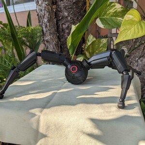 Pode incluir: Um dispositivo mec&acirc;nico preto, semelhante a uma aranha, com pernas articuladas e um corpo central com um detalhe circular vermelho. O dispositivo est&aacute; sobre uma superf&iacute;cie clara, com vegeta&ccedil;&atilde;o ao fundo.