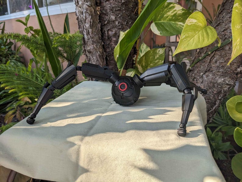 Pode incluir: Um dispositivo mec&acirc;nico preto, semelhante a uma aranha, com pernas articuladas e um corpo central com um detalhe circular vermelho. O dispositivo est&aacute; sobre uma superf&iacute;cie clara, com vegeta&ccedil;&atilde;o ao fundo.