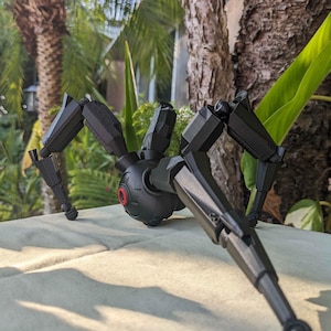 Pode incluir: Um brinquedo de aranha mec&acirc;nica preta com pernas articuladas e um corpo central. O brinquedo tem um detalhe vermelho semelhante a um olho e est&aacute; posicionado em uma superf&iacute;cie de cor clara, com um fundo natural desfocado de &aacute;rvores e folhagens.