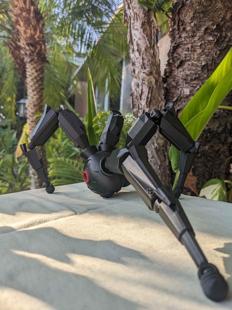 Pode incluir: Um brinquedo de aranha mec&acirc;nica preta com pernas articuladas e um corpo central. O brinquedo tem um detalhe vermelho semelhante a um olho e est&aacute; posicionado em uma superf&iacute;cie de cor clara, com um fundo natural desfocado de &aacute;rvores e folhagens.