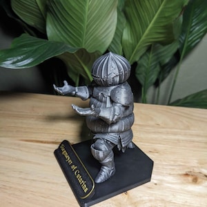 May include: A silver-colored figurine of a knight in armor, posed with arms outstretched. The figurine is mounted on a black base with the text "Siegmeyer of Catarina" in gold lettering. The knight's helmet has a segmented design.