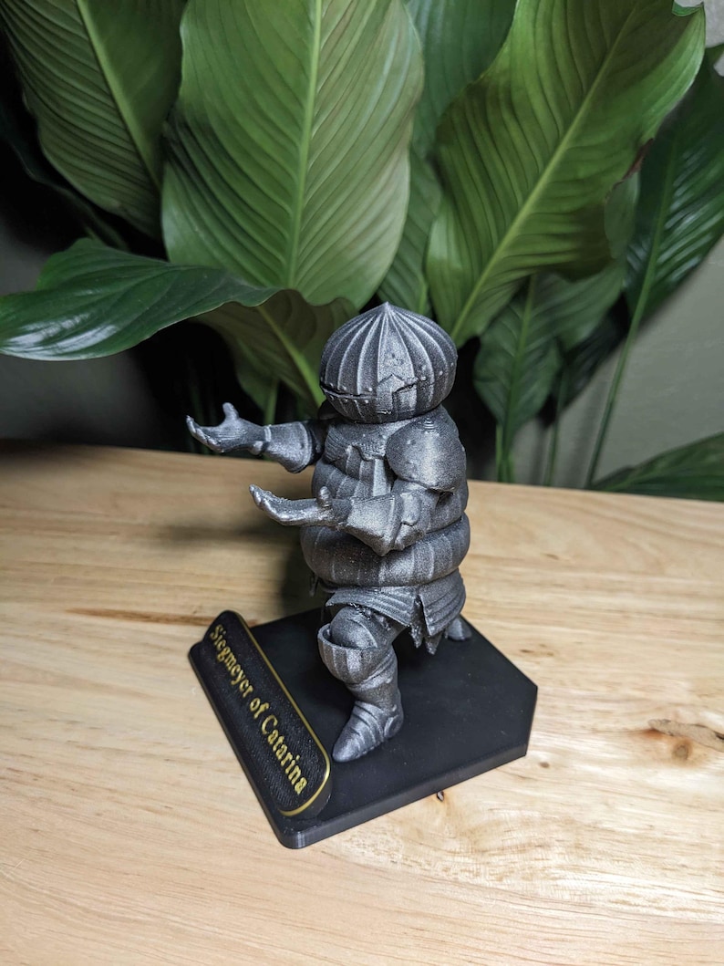 May include: A silver-colored figurine of a knight in armor, posed with arms outstretched. The figurine is mounted on a black base with the text "Siegmeyer of Catarina" in gold lettering. The knight's helmet has a segmented design.
