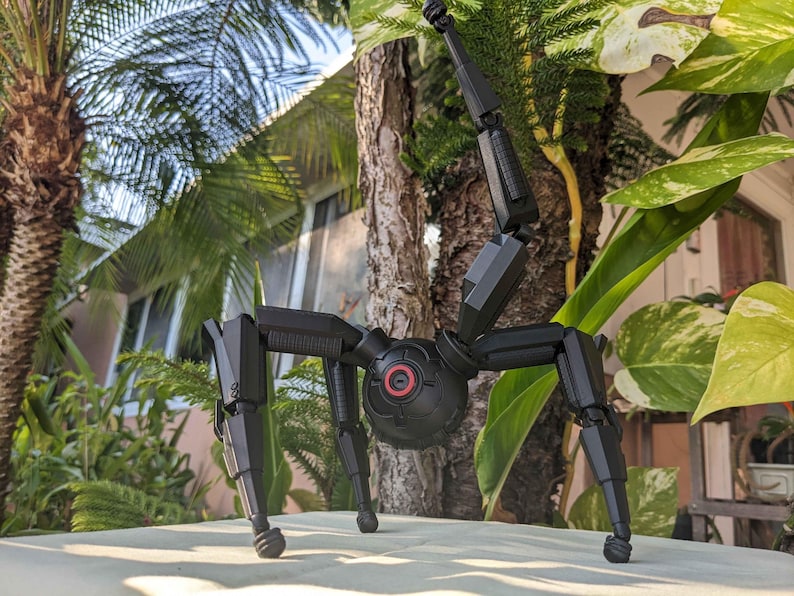 Pode incluir: Um brinquedo rob&oacute;tico preto, mec&acirc;nico, semelhante a uma aranha, com pernas articuladas e um corpo esf&eacute;rico central. O rob&ocirc; tem um detalhe circular vermelho no corpo e est&aacute; posicionado sobre uma superf&iacute;cie de cor clara, com um fundo natural de &aacute;rvores e folhagem.