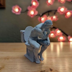 Könnte beinhalten: Eine hellgraue, gesprenkelte Figur, die eine sitzende Person in kontemplativer Haltung darstellt, das Kinn auf eine Hand gestützt, während sie auf einer kleinen Toilette sitzt. Die Skulptur hat eine matte, texturierte Oberfläche in einer Farbe.