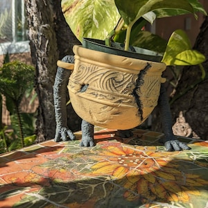 May include: A unique, tan-colored planter with a cracked design, supported by gray, creature-like legs and arms. The planter holds a green plant with large leaves. The planter sits on a colorful mosaic surface with a sunflower design.