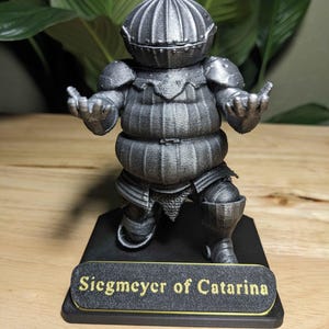 May include: A detailed, gray figurine of a knight in armor, with the text "Siegmeyer of Catarina" on a base. The knight has a round helmet, shoulder pads, and a segmented torso. The figurine is posed with arms outstretched.