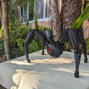 Pode incluir: Um brinquedo de aranha mec&acirc;nica preta com pernas articuladas e um corpo central com um detalhe circular vermelho. O brinquedo est&aacute; posicionado em uma superf&iacute;cie de cor clara, com um fundo natural ao ar livre de &aacute;rvores e folhagem.