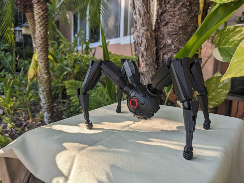 Pode incluir: Um brinquedo de aranha mec&acirc;nica preta com pernas articuladas e um corpo central com um detalhe circular vermelho. O brinquedo est&aacute; posicionado em uma superf&iacute;cie de cor clara, com um fundo natural ao ar livre de &aacute;rvores e folhagem.