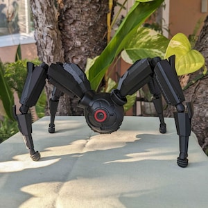 May include: A black and grey mechanical spider toy with articulated legs and a central body featuring a red circular detail. The toy is positioned on a light-coloured surface, with greenery in the background.