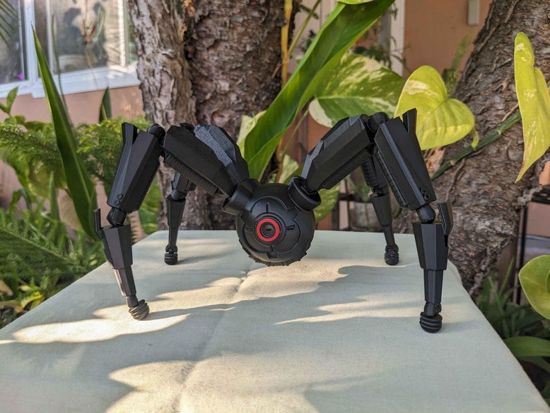 Pode incluir: Um brinquedo de aranha mec&acirc;nica preta e cinza com pernas articuladas e um corpo central com um detalhe circular vermelho. O brinquedo est&aacute; posicionado em uma superf&iacute;cie de cor clara, com vegeta&ccedil;&atilde;o ao fundo.