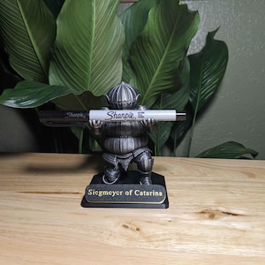 May include: A decorative figurine of a knight holding a Sharpie marker. The figurine is detailed with armor and a helmet, and the base reads "Siegmeyer of Catarina". The Sharpie is black and white.