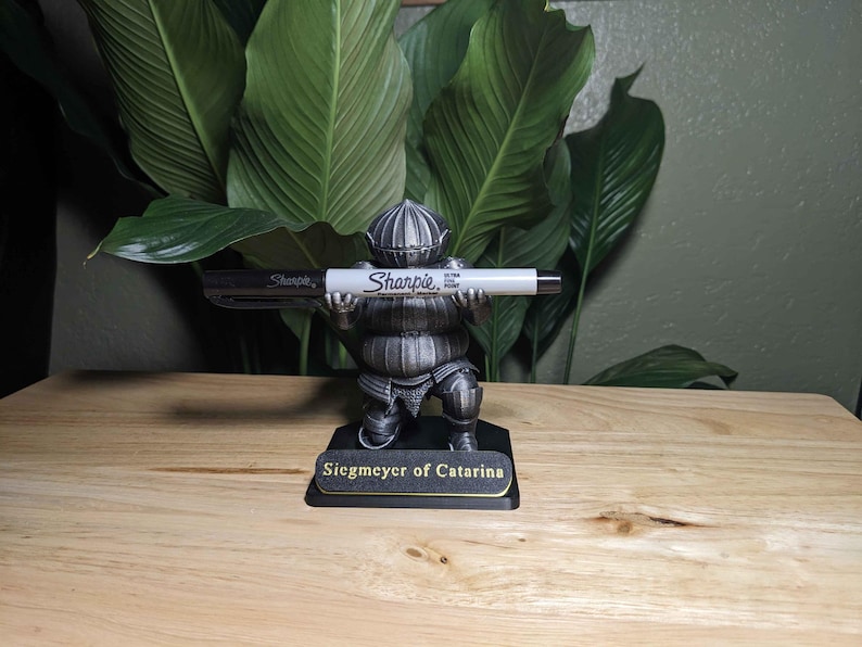 May include: A decorative figurine of a knight holding a Sharpie marker. The figurine is detailed with armor and a helmet, and the base reads "Siegmeyer of Catarina". The Sharpie is black and white.