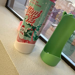 May include: A teal and red can of "Mani Na" energy drink with a winter wonderland theme, featuring a snowman and palm trees. The can is in a white and pink holder, next to a green holder.