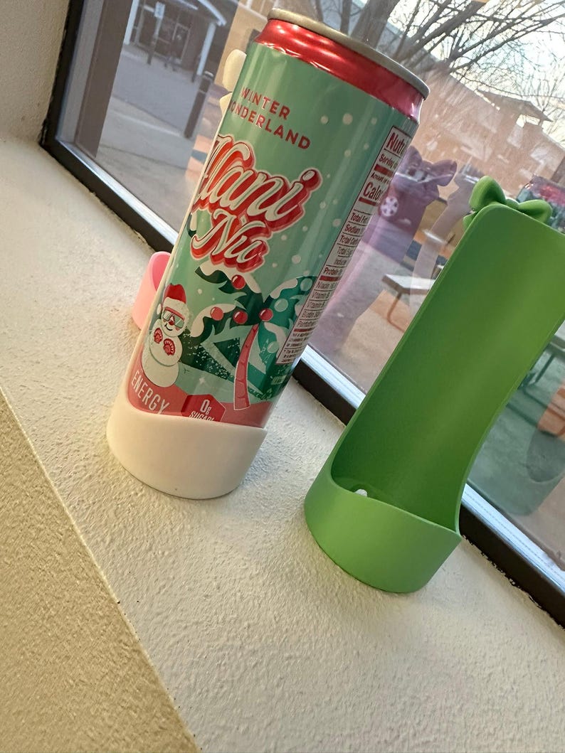 May include: A teal and red can of "Mani Na" energy drink with a winter wonderland theme, featuring a snowman and palm trees. The can is in a white and pink holder, next to a green holder.