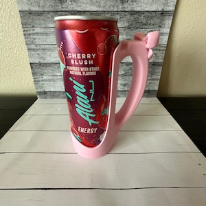 May include: A pink 3D-printed handle designed to hold a can. The handle is shaped like a mug with a bow detail. A red can of "Cherry Slush" energy drink is held in the handle.