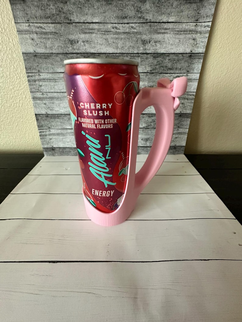 May include: A pink 3D-printed handle designed to hold a can. The handle is shaped like a mug with a bow detail. A red can of "Cherry Slush" energy drink is held in the handle.