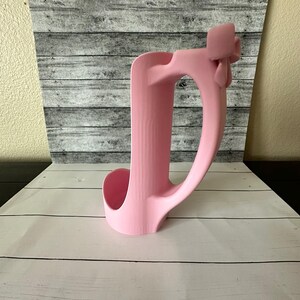 May include: A pink, molded plastic cup holder with a handle. The holder has a curved design with a circular opening for the cup and a handle for carrying. The background is a gray and white wood-like pattern.