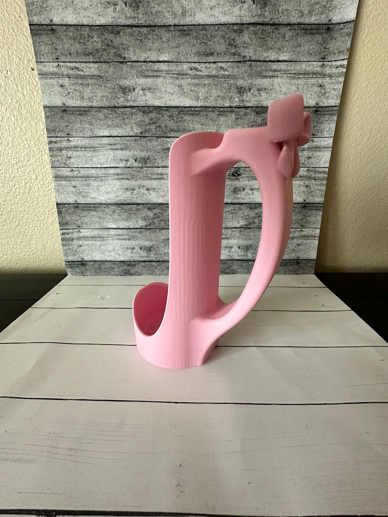 May include: A pink, molded plastic cup holder with a handle. The holder has a curved design with a circular opening for the cup and a handle for carrying. The background is a gray and white wood-like pattern.