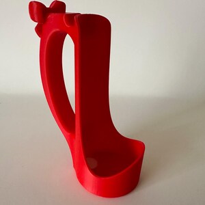 May include: A bright red, 3D-printed phone holder. The holder features a curved handle and a bow-shaped detail at the top. The base is a rounded rectangle, designed to hold a phone. The holder is made of a smooth, solid material.