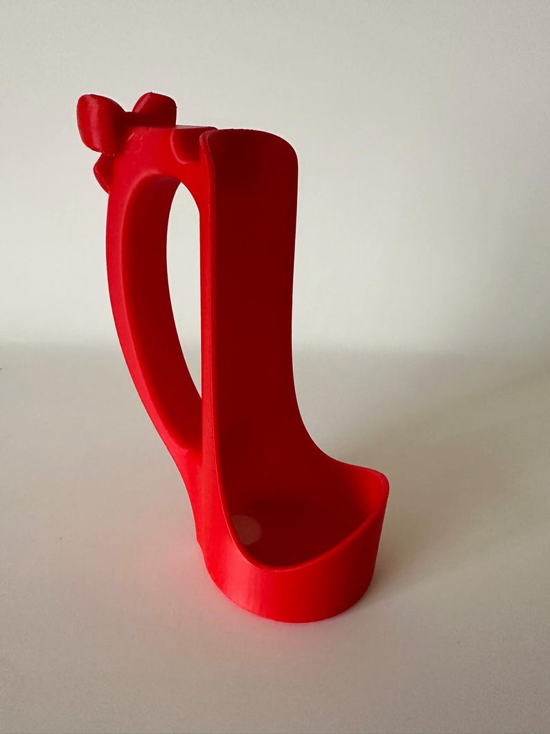 May include: A bright red, 3D-printed phone holder. The holder features a curved handle and a bow-shaped detail at the top. The base is a rounded rectangle, designed to hold a phone. The holder is made of a smooth, solid material.