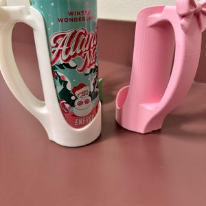 May include: Two 3D-printed can holders. One is white and holds a can with a teal and red label that reads "Winter Wonderland Allan N's Energy". The other is pink with a bow detail.