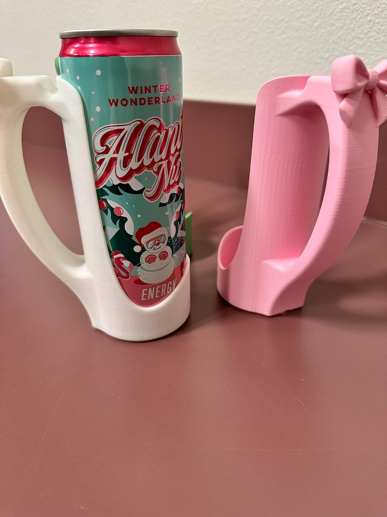 May include: Two 3D-printed can holders. One is white and holds a can with a teal and red label that reads "Winter Wonderland Allan N's Energy". The other is pink with a bow detail.