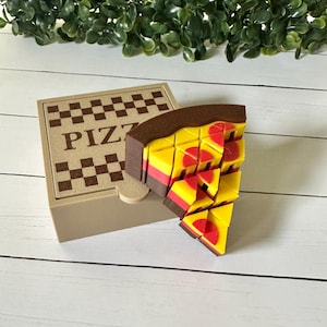 May include: A tan pizza box with the word "PIZZA" printed on the lid. A slice of pizza is next to the box, made of yellow, red, and brown pieces. The pizza slice is a puzzle.